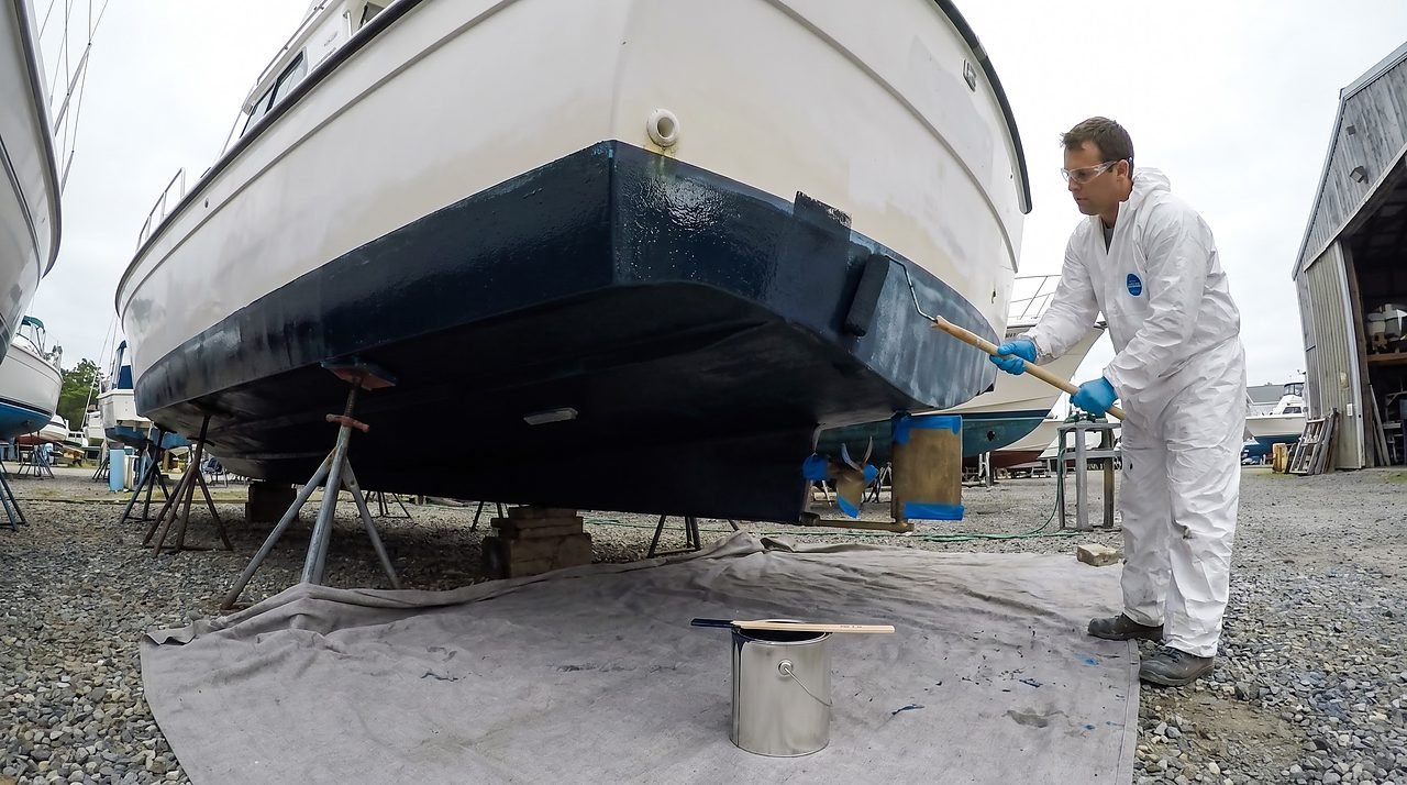 A person applying dark antifouling bottom paint to a hauled-out fiberglass powerboat hull