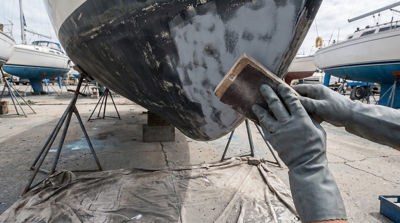 gloved hands sanding boat hull bottom with 80-grit sandpaper in a boatyard
