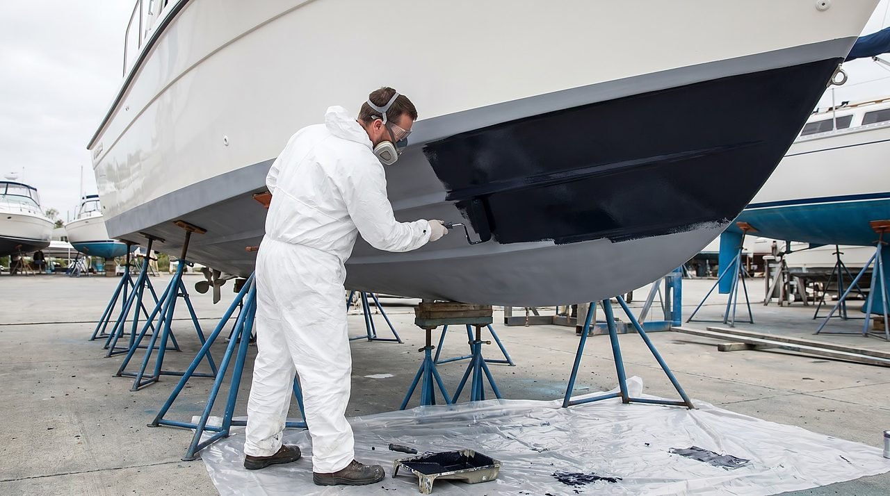 person in coveralls applying dark antifouling paint to a fiberglass boat hull with a roller